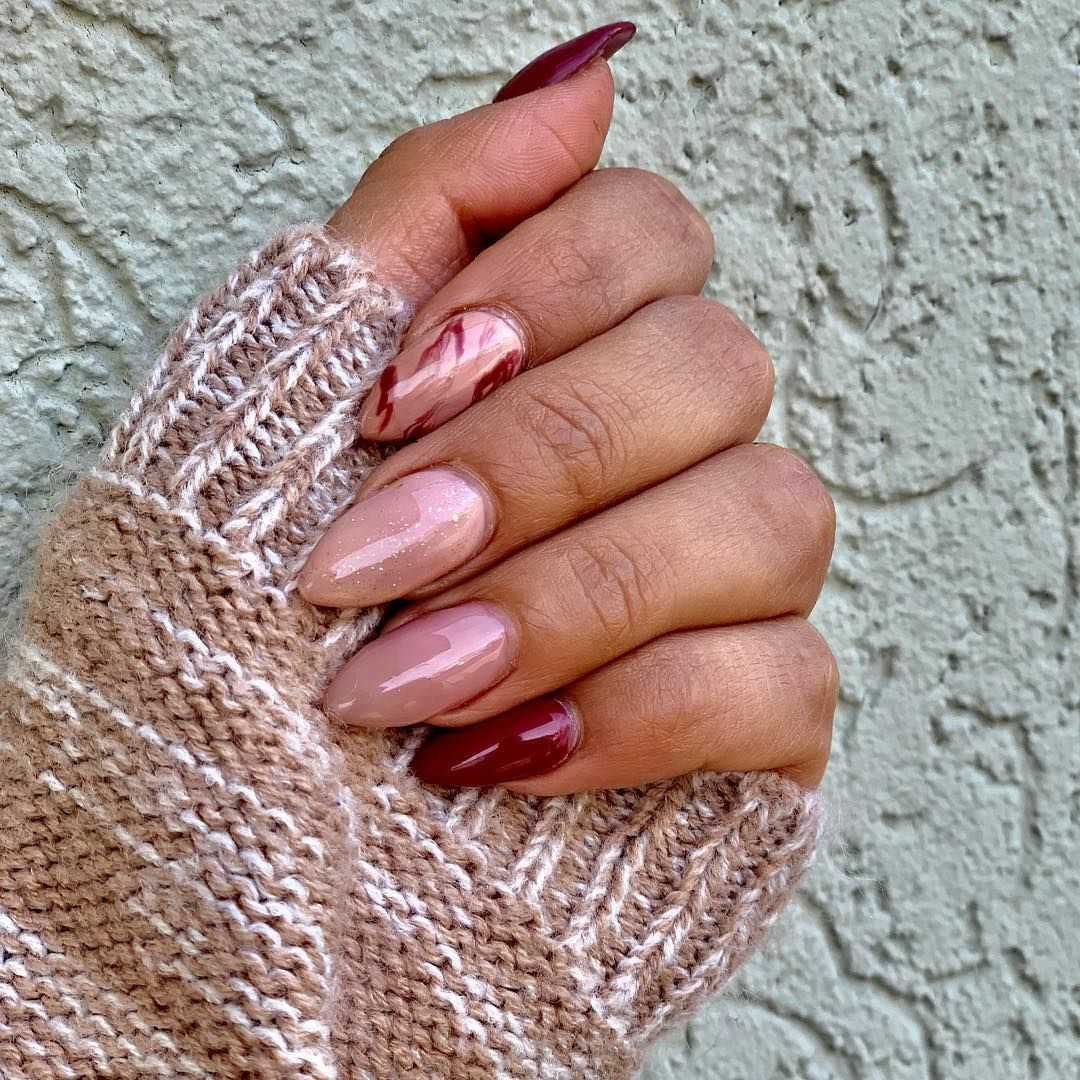 Hand with maroon and pink nail art, in a striped beige sweater sleeve against a textured wall.